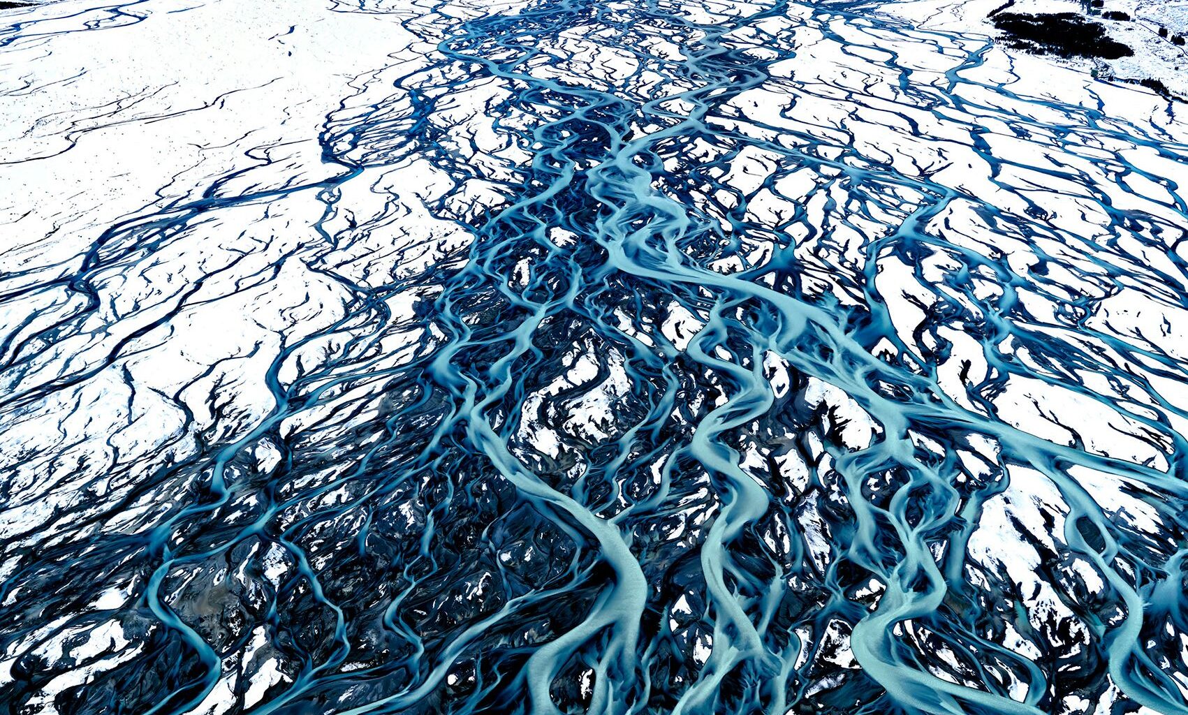 Aerial view of a glacial river meandering through a snowy landscape, surrounded by towering mountains and blue sky
