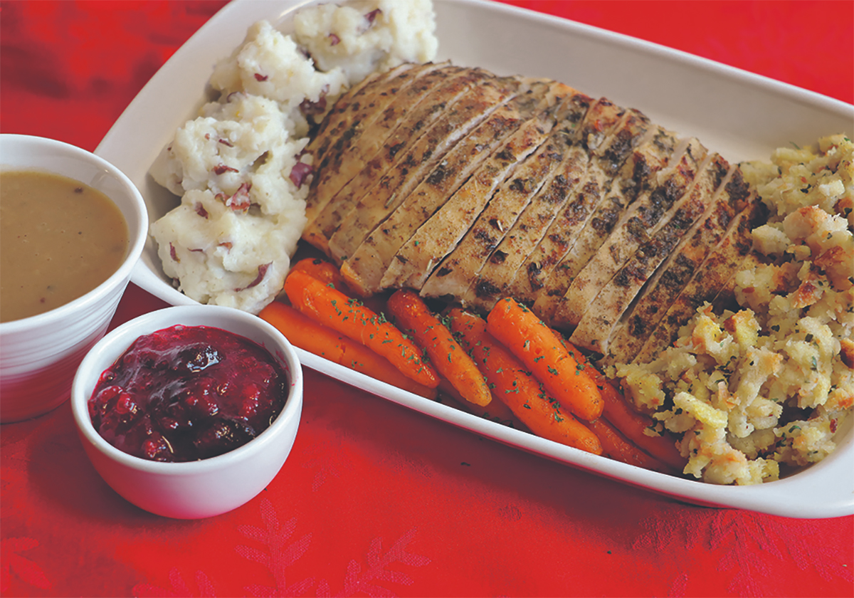 Slices of roasted turkey, mashed potatoes, carrots and stuffing sit on a serving plate with a bowl of gravy and another smaller bowl of cranberry sauce next to it.