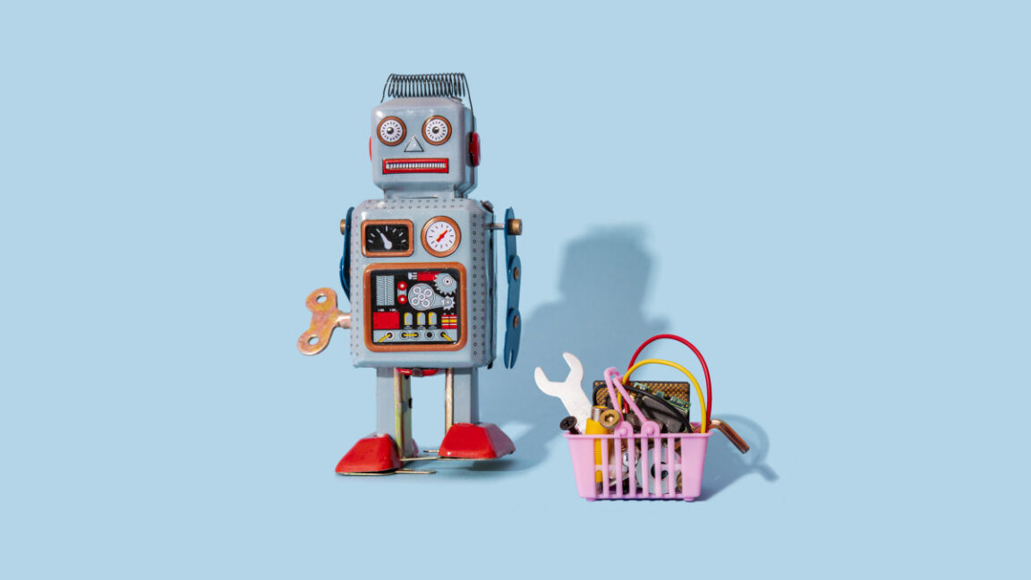 Studio shot of vintage robot toy standing by miniature shopping basket filled with electronic equipment