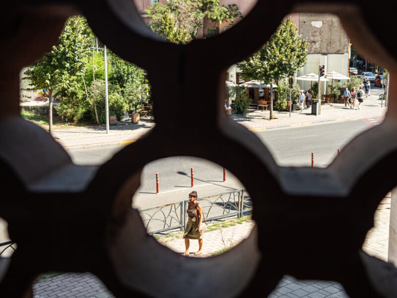 A woman walks on a sunny sidewalk, seen through a decorative stone lattice, with a street, trees, and people at an outdoor cafe visible in the background.