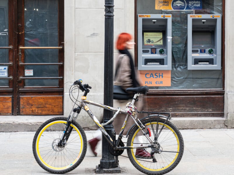 A bicycle is locked to a lamppost on a city sidewalk. A person with red hair walks by, blurred in motion, wearing a mask. Two ATMs are mounted on the wall behind them.