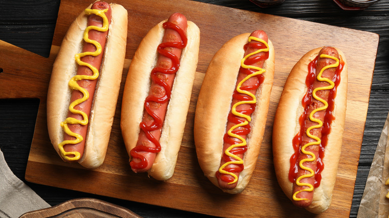 Four hot dogs topped with mustard, ketchup, and both on a wooden serving board