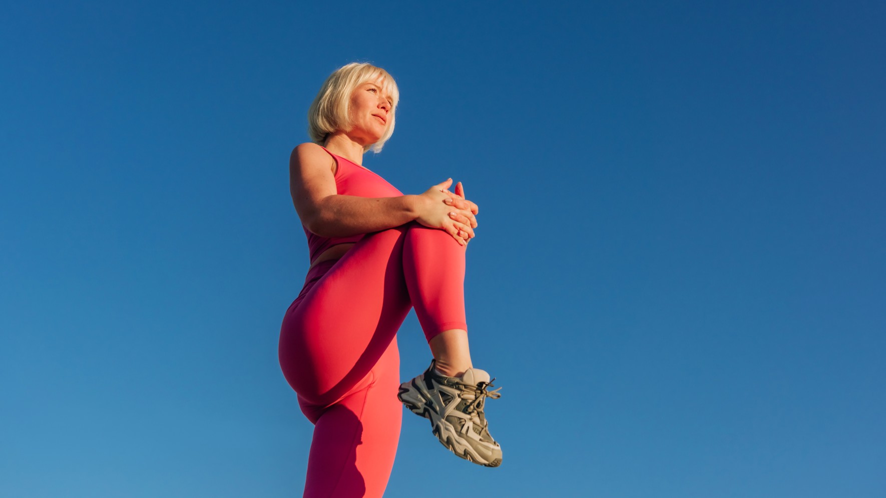 Blonde woman in pink workout gear against a blue sky backdrop holding her knee to her chest for a stretch