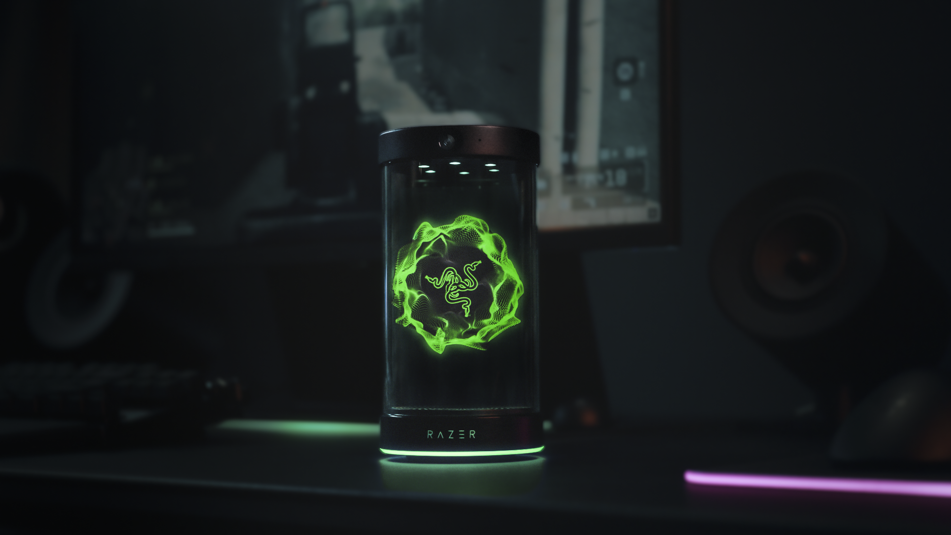 The Project AVA desk companion from Razer sits atop a black desk. A holographic image of Razer's logo lights up the screen in green.