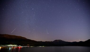 White streaks of meteors are seen against a dark purple night sky with a frozen lake with lights below pushed against silhouetted mountains in the background