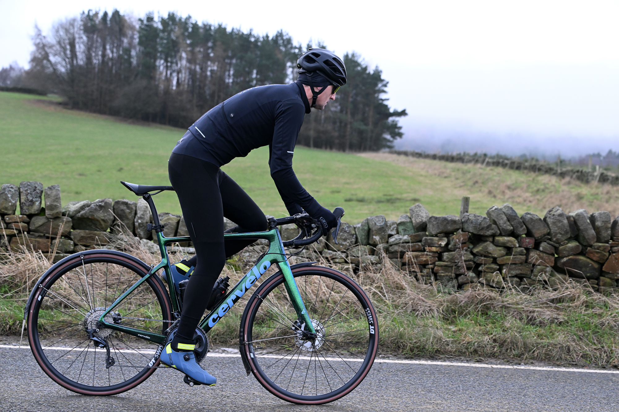 Side view of a man riding out of the saddle wearing a black jacket, black tights and a black helmet on a green Cervelo bike