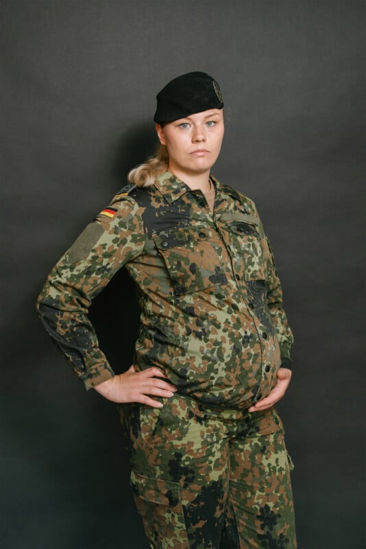 A pregnant woman in German military camouflage uniform and a black beret stands with one hand on her hip and the other on her belly, against a plain dark background.