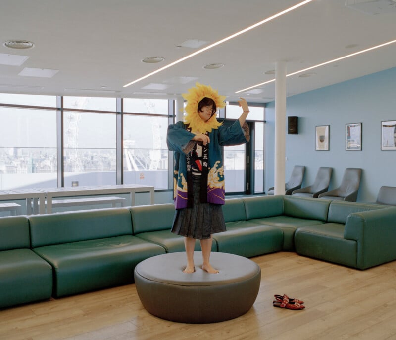 A person wearing a sunflower costume and colorful robe stands barefoot on a round ottoman in a modern lounge area. Their sandals are on the floor nearby, and the room features green couches and large windows.