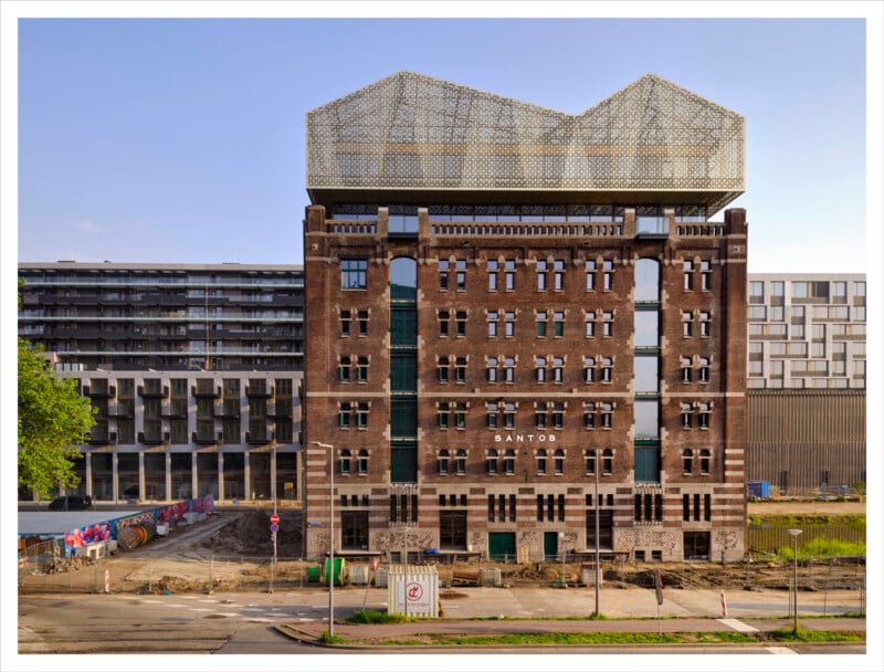 A tall, brick building with "Santos" written on the front stands amid construction work. The upper floors have a modern, geometric metal structure. Nearby, contemporary buildings and a street are visible.