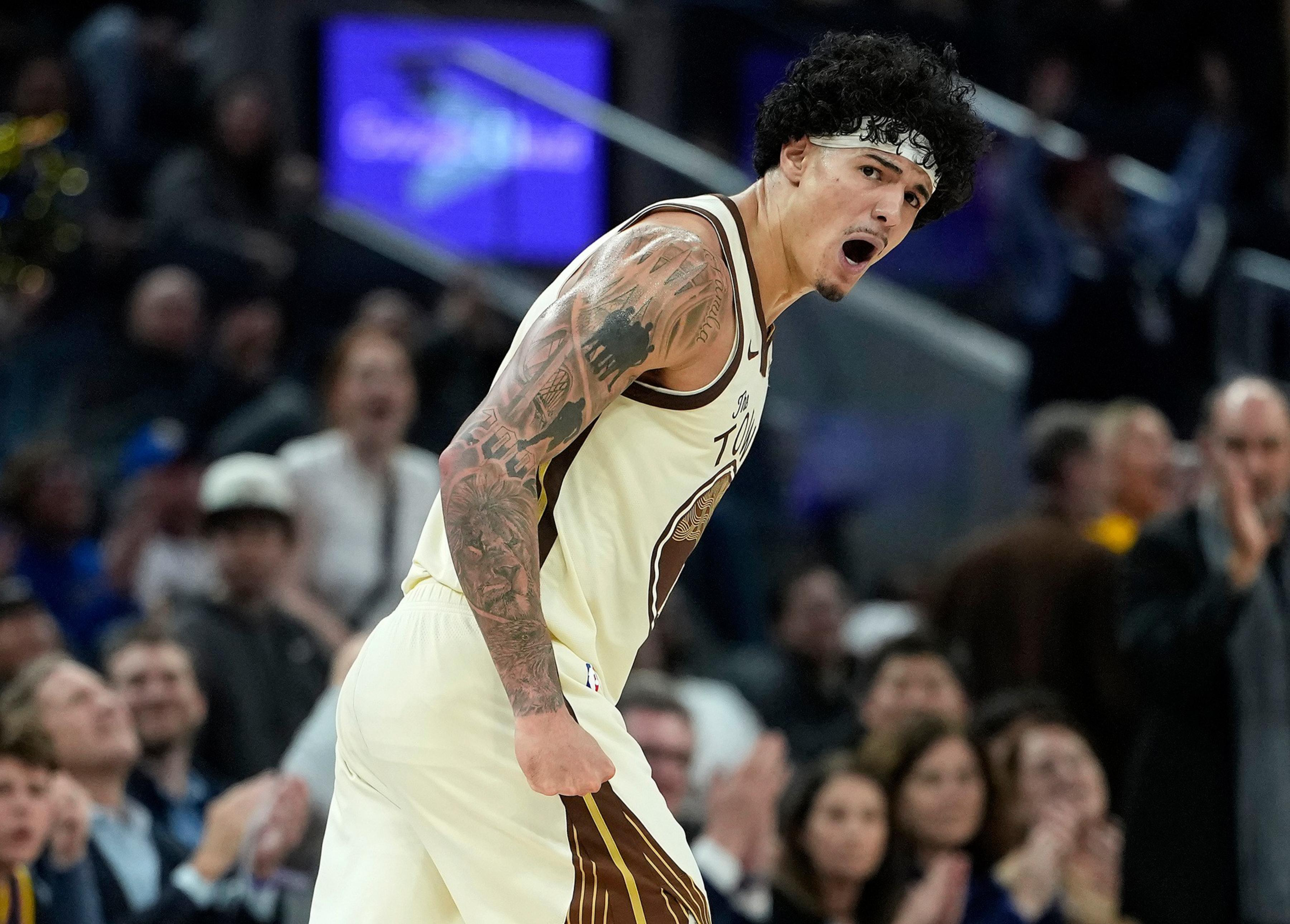 A basketball player with tattoos and a headband is shouting passionately during a game, with a crowd blurred in the background.
