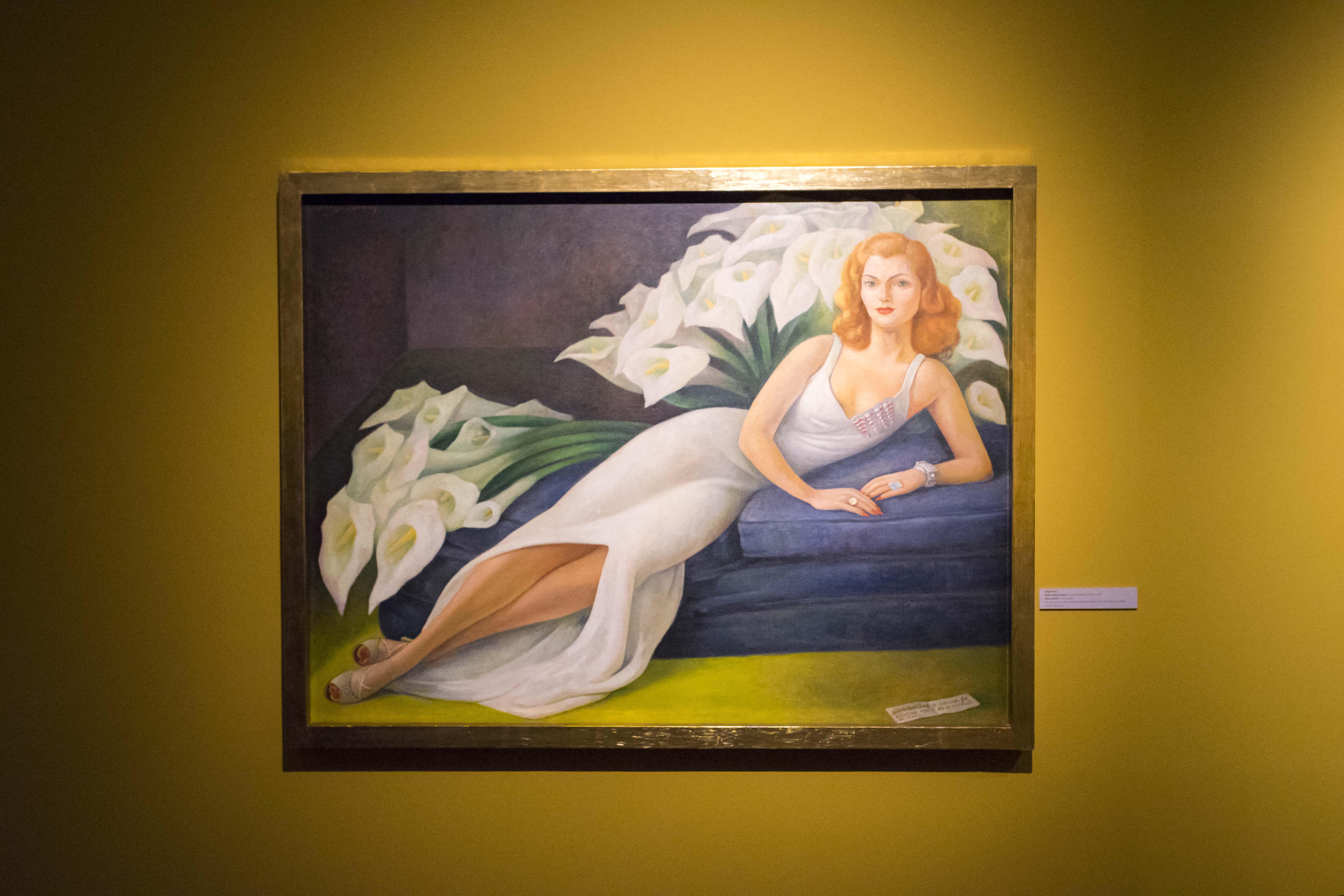 Illustration of Diego Rivera's "Portrait of Natasha Gelman" on display, featuring a woman in a white dress lying on a blue cushion with white calla lilies behind her.