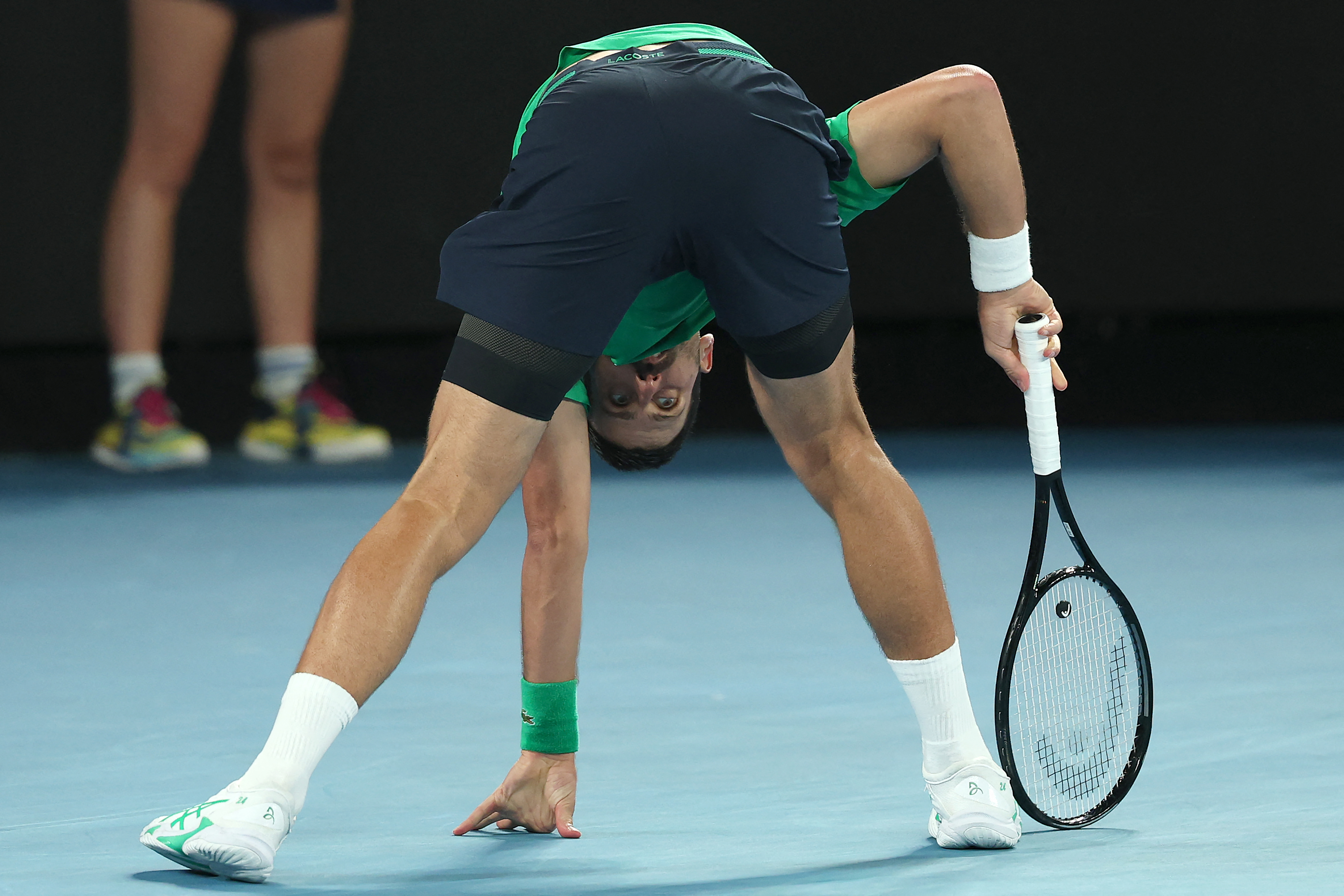 Novak Djokovic bending over with a tennis racket, looking up towards the camera.