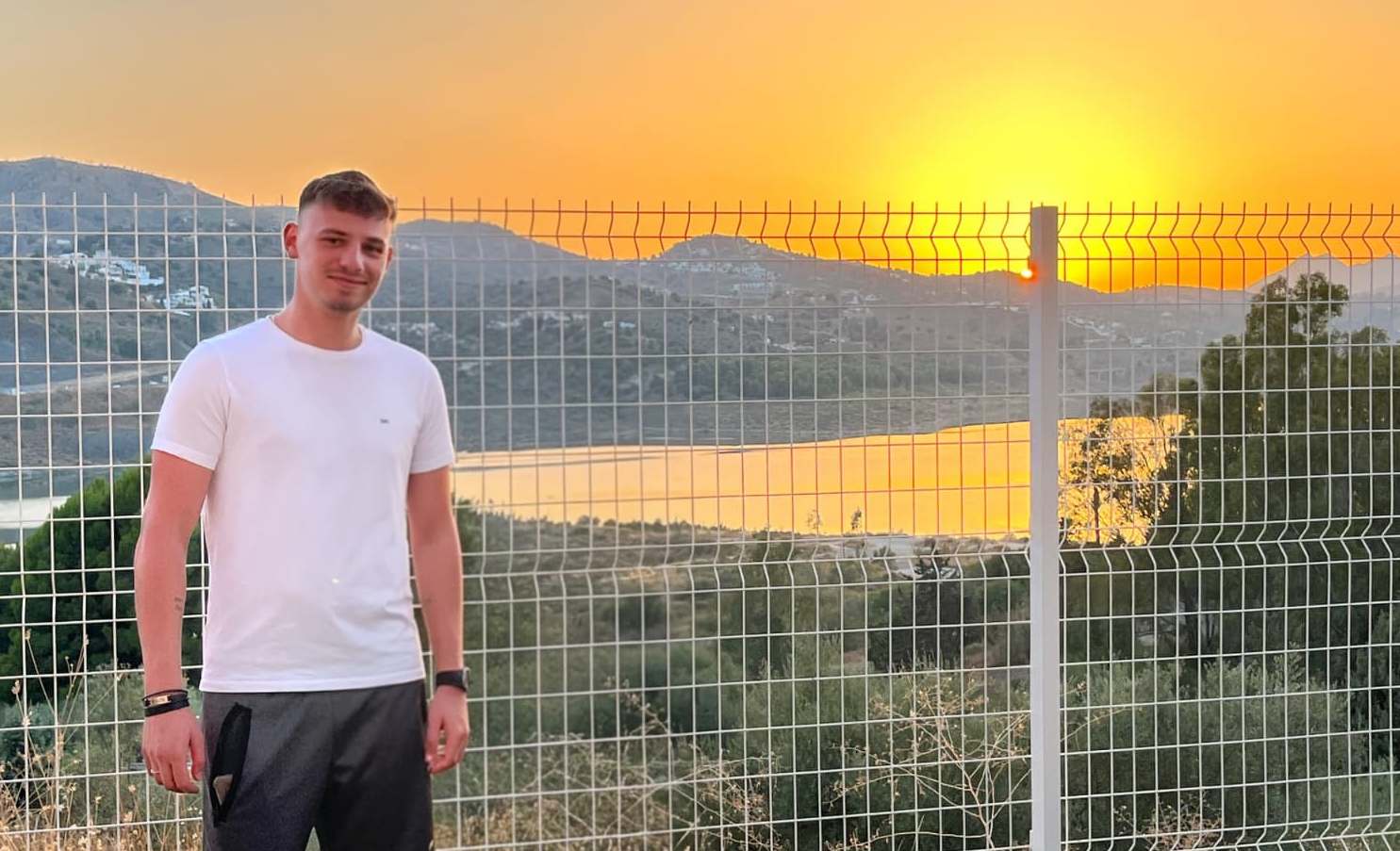 A man stands in front of a wire fence with a sunset over mountains and a body of water behind him.