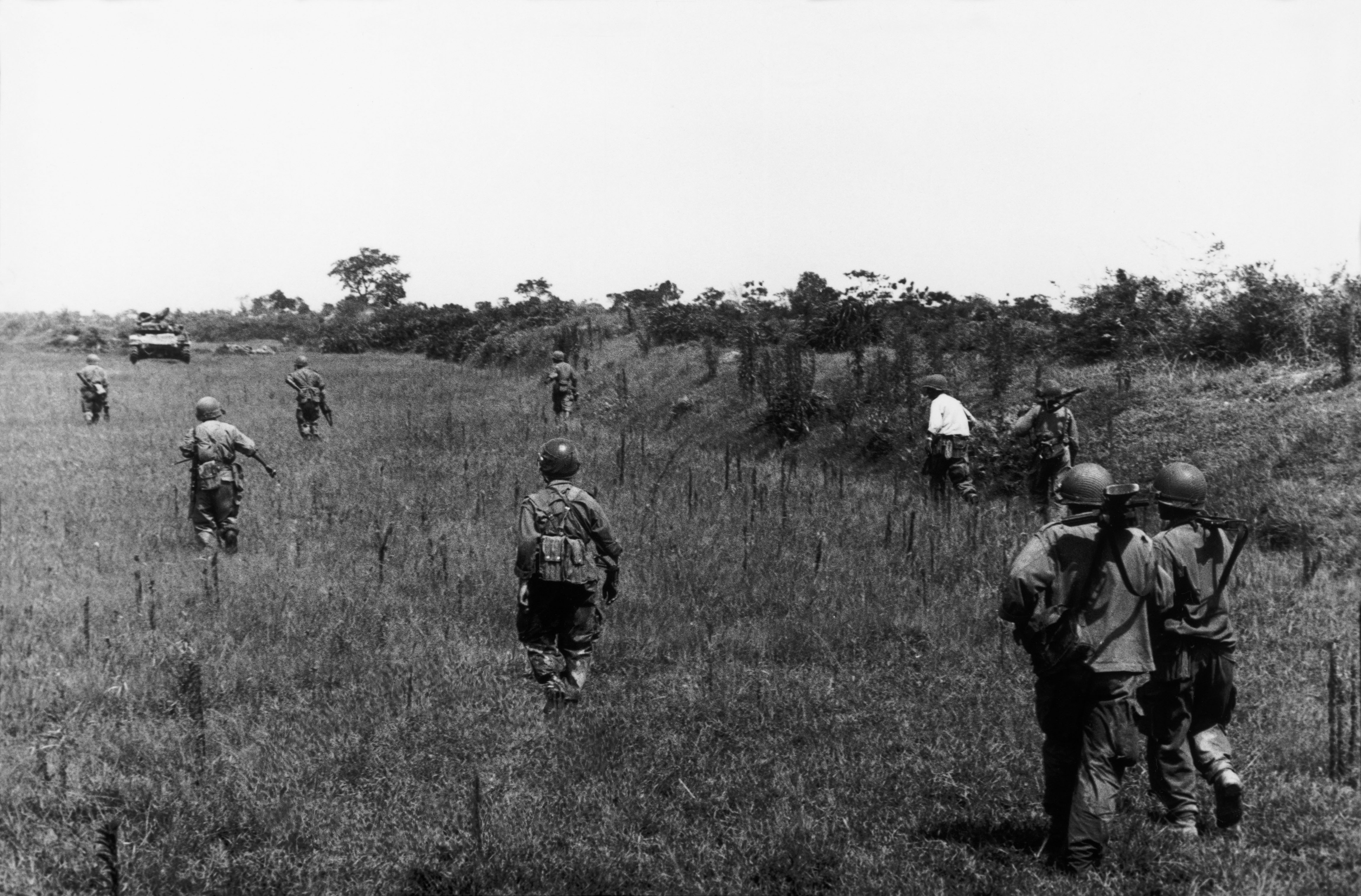 Soldiers walking through a field with a tank in the background.