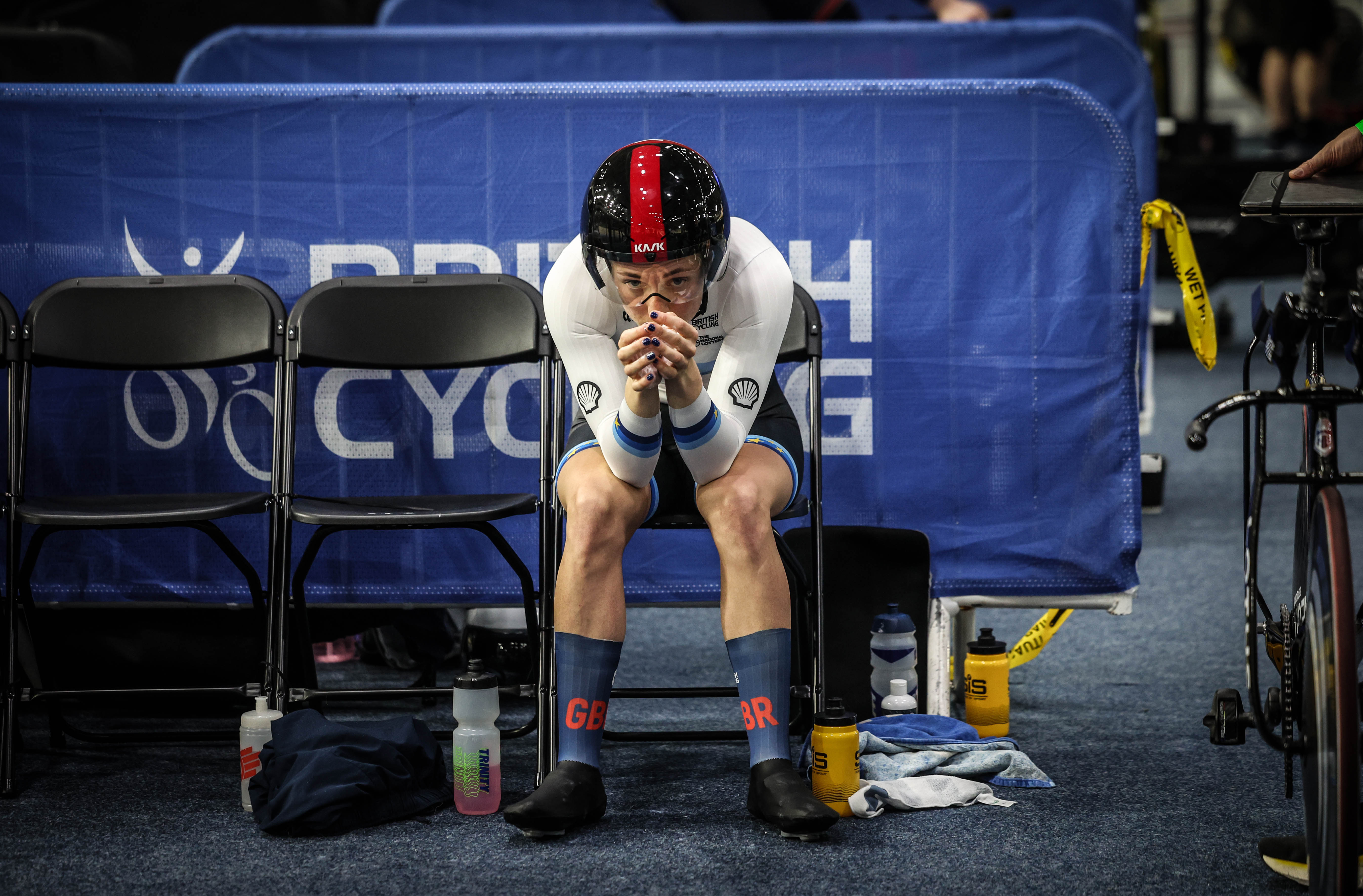 A cyclist in full gear, wearing a helmet and a white and blue uniform, sits on a black folding chair, hands clasped together, looking down.