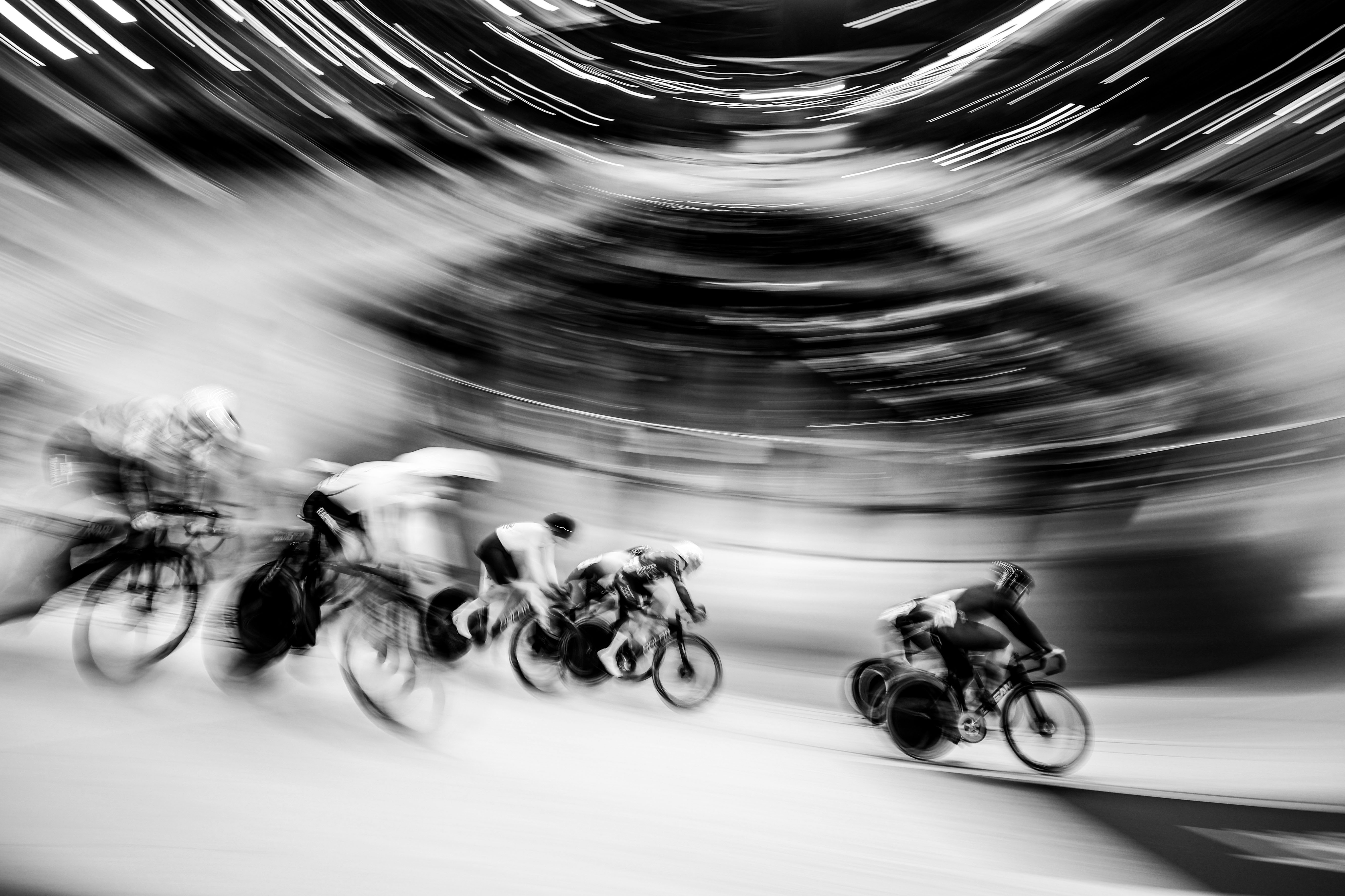 Black and white photograph capturing track cyclists racing at Manchester Velodrome with motion blur to convey speed.