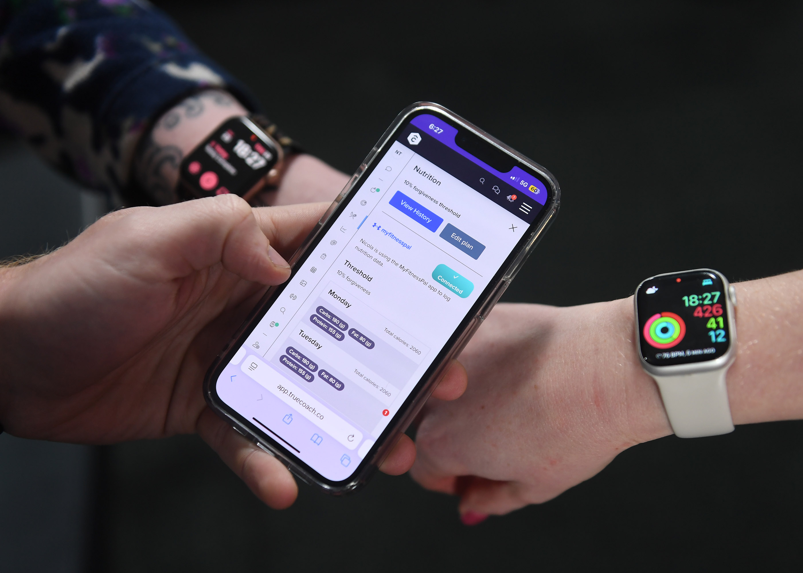 A person holds a phone displaying the TrueCoach app showing nutrition data, while another person's wrist wears an Apple Watch showing heart rate and activity.
