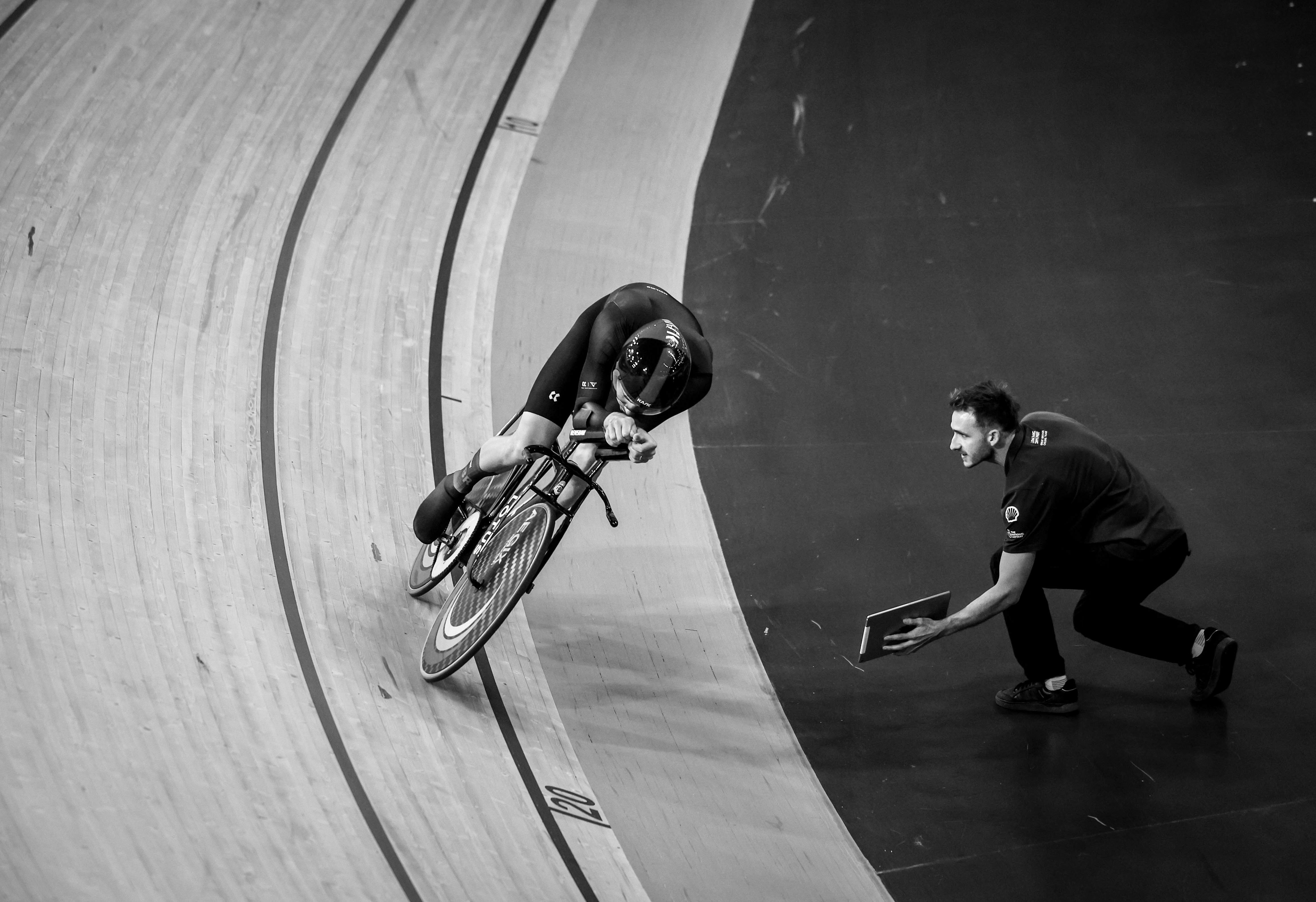 A cyclist in full aerodynamic gear on a velodrome track with a man kneeling beside the track holding a tablet.