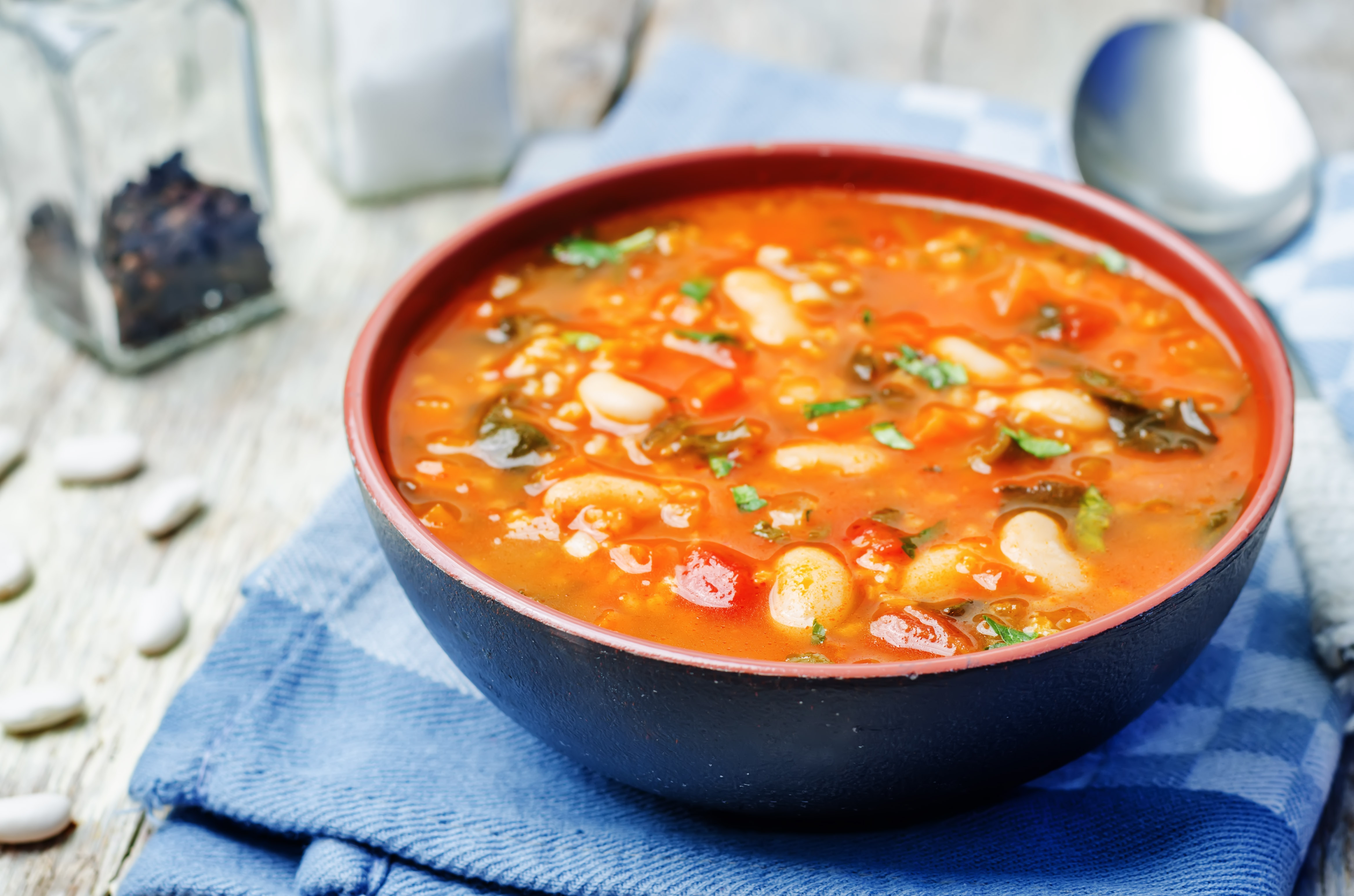 A bowl of tomatoes, spinach, white beans, and millet soup on a blue checkered napkin.