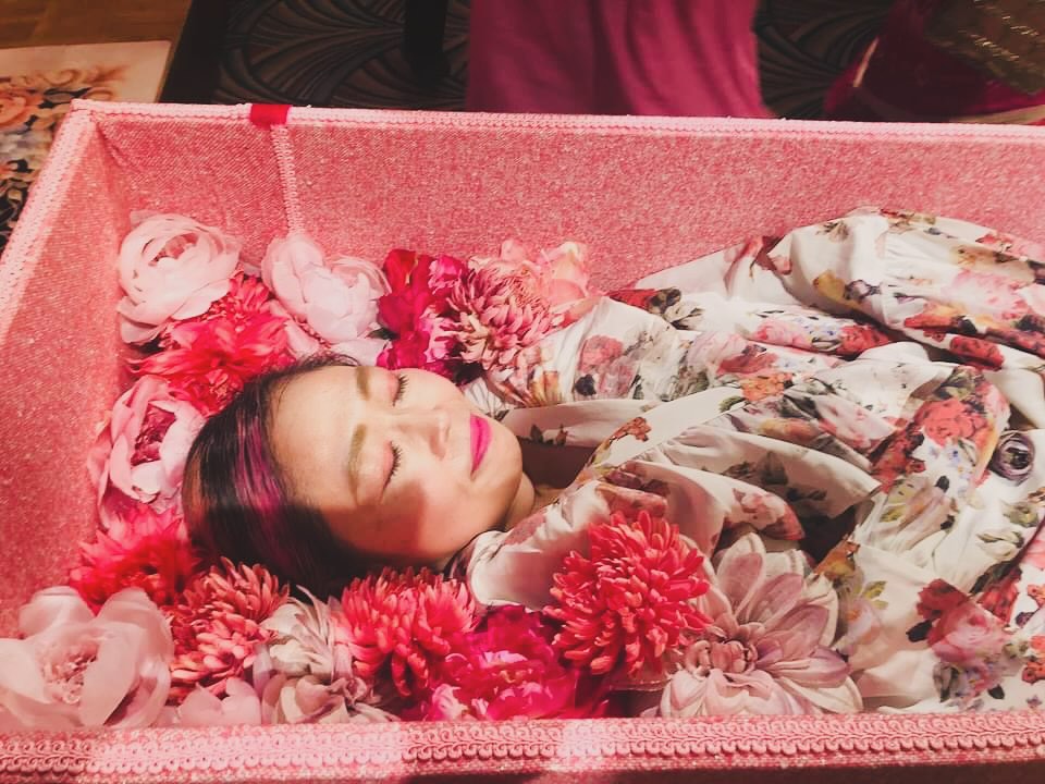 A person lying in a pink coffin surrounded by pink flowers.