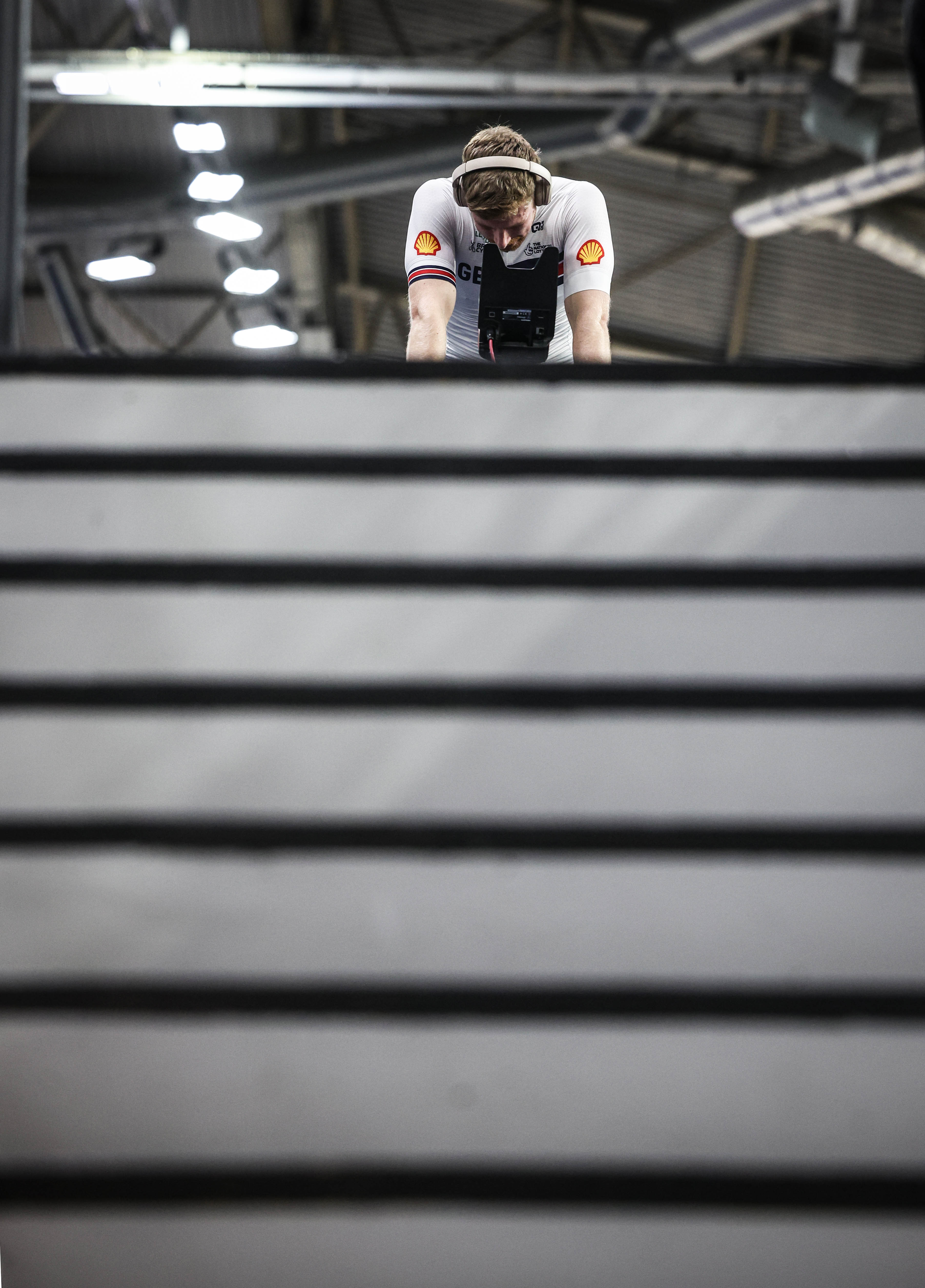 A cyclist in a white jersey with sponsor logos and headphones, bending over handlebars.