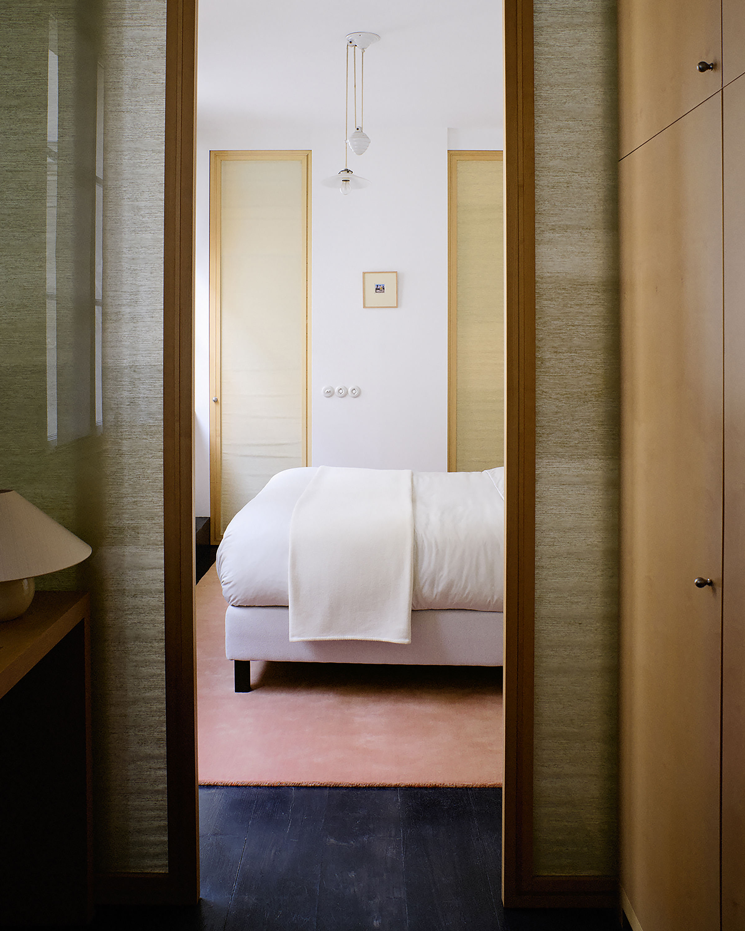 A bedroom with a bed with white bedding, a pink rug, and dark wooden floors, seen through an open doorway.