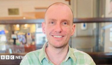 Ben Howard, a man with a buzz cut and stubble, is pictured smiling at the camera. He is wearing a light green, checkered shirt with a collar. He appears to be sat in a pub.