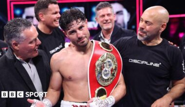 Josh Kelly holds up his hand and has the IBF light-middleweight title over his shoulder as he stands next to people in a boxing ring