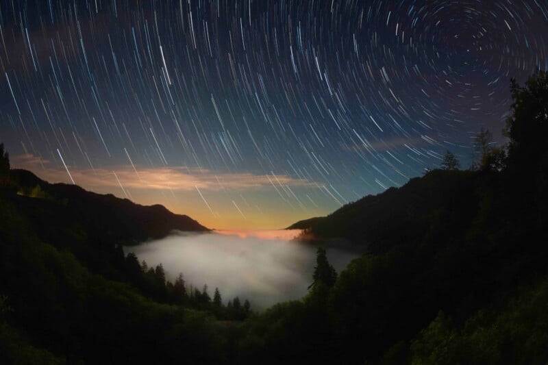 A night sky with circular star trails above a misty valley surrounded by dark, forested hills. The horizon glows faintly orange, hinting at dawn or sunset, while trees are silhouetted against the low-lying fog.