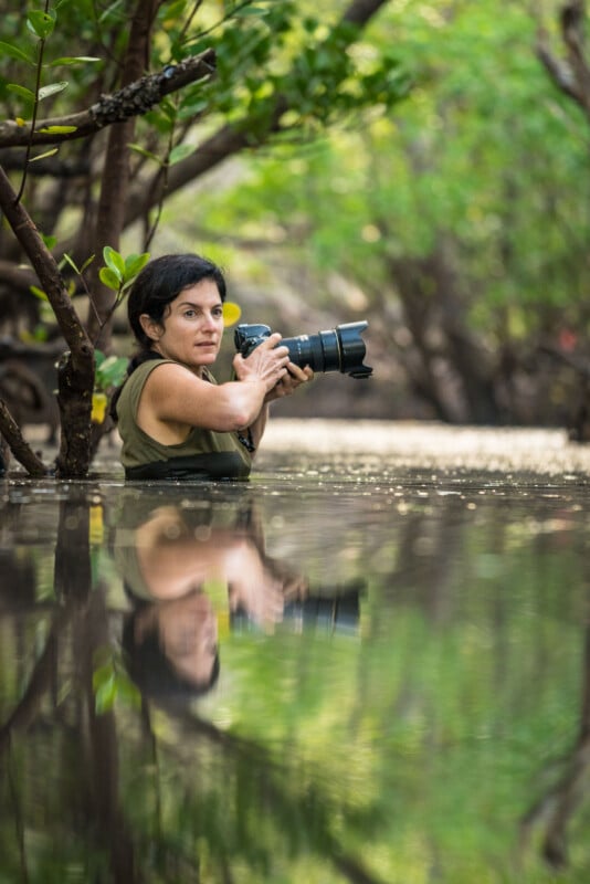 A woman stands waist-deep in water among trees, holding a camera with a long lens, surrounded by lush green foliage, and her reflection is visible on the water’s surface.