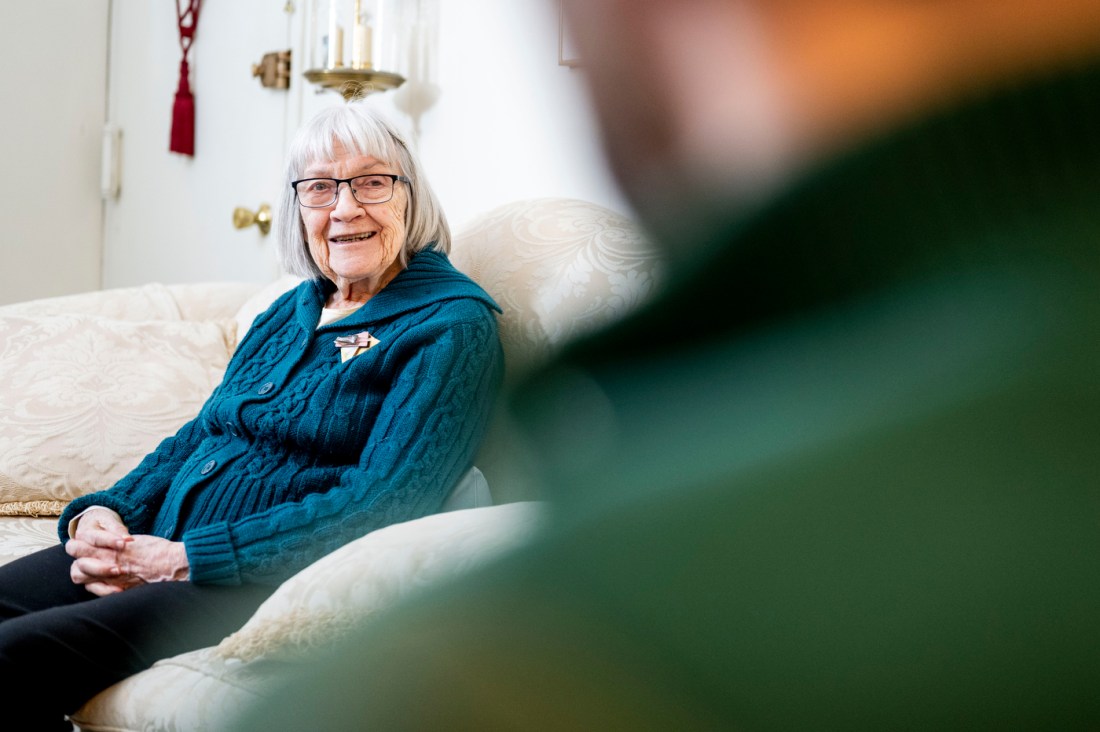 Ruth Gove in her home, smiling on a white sofa.