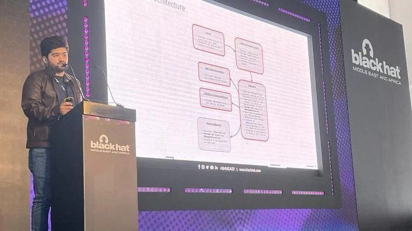 Etizaz Mohsin standing at the podium giving a presentation. He has black hair and wearing a brown leather jacket and jeans