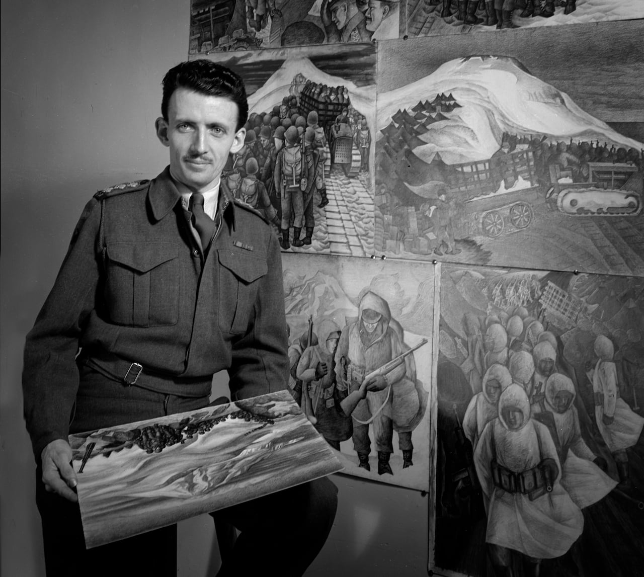 Black and white photo from 1945. A man in military uniform, the artist E.J. Hughes, stands next to several paintings of soldiers. He holds another.