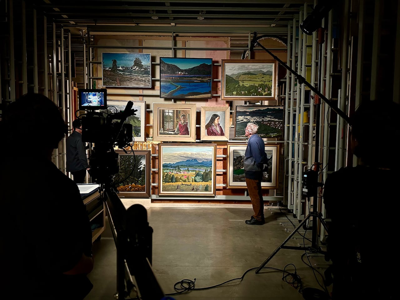 A film crew, in shadow, records a man standing in front of a wall of paintings. They are inside a gallery vault.