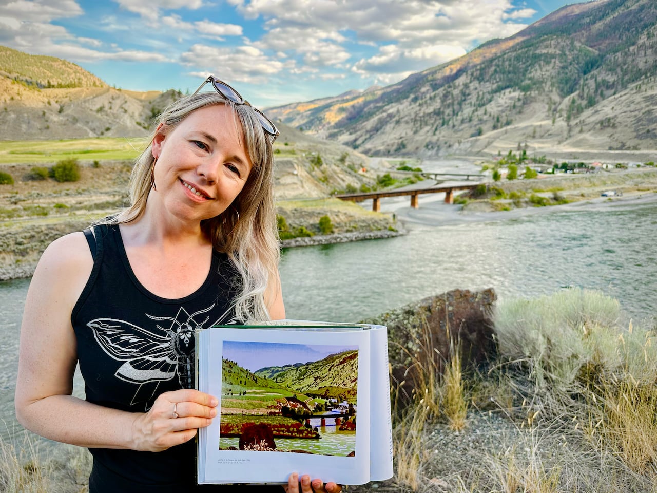 A woman with blonde hair holds a print of a landscape painting. The real-life landscape which inspired it rises behind her.