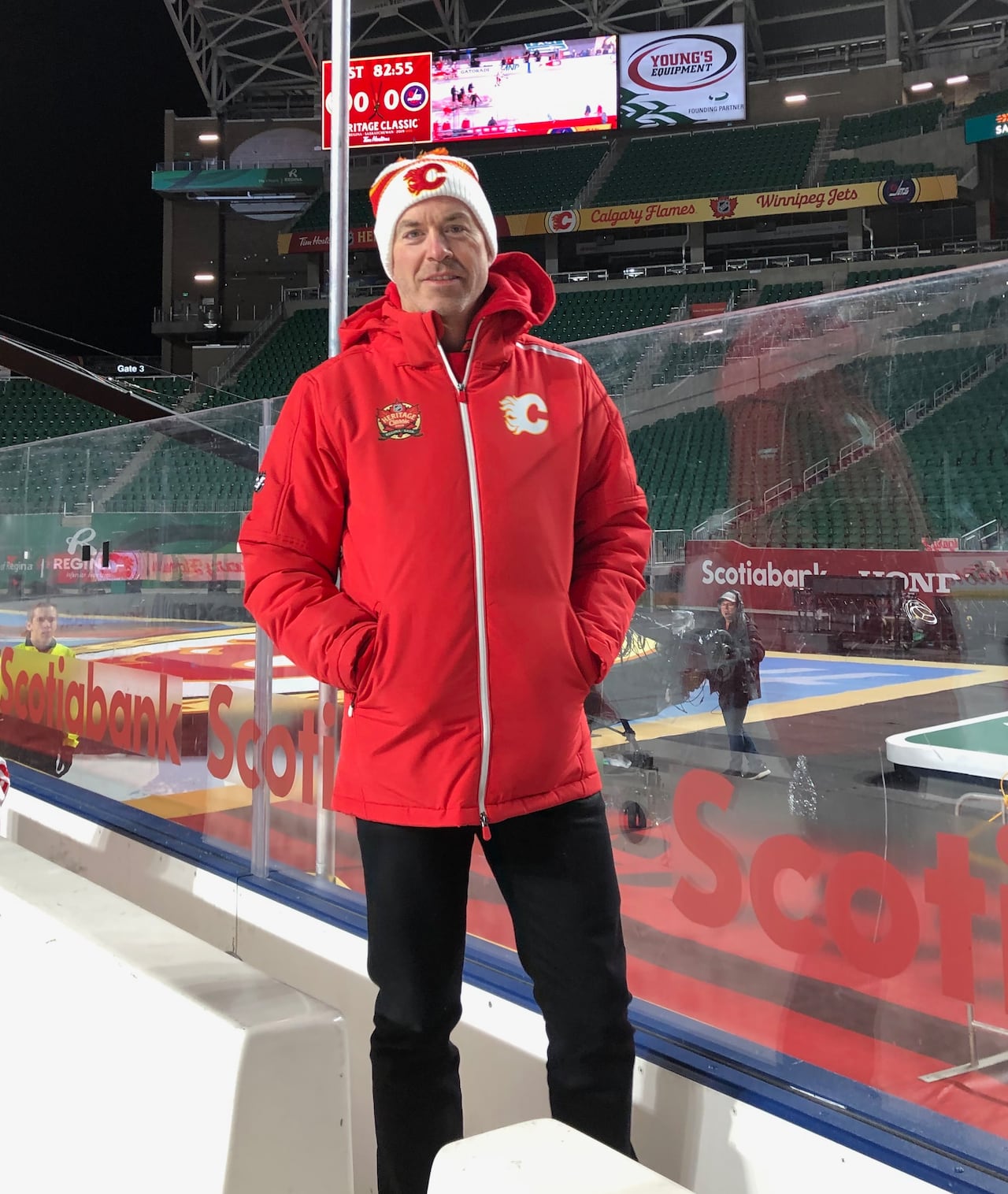 A man wearing a red jacket and toque stands on a hockey bench