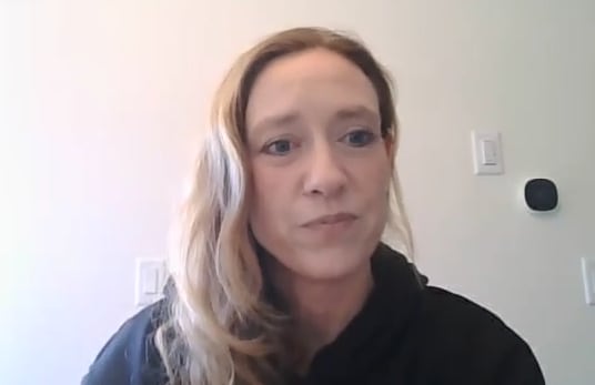 A woman with blond hair sits in front of a white wall.