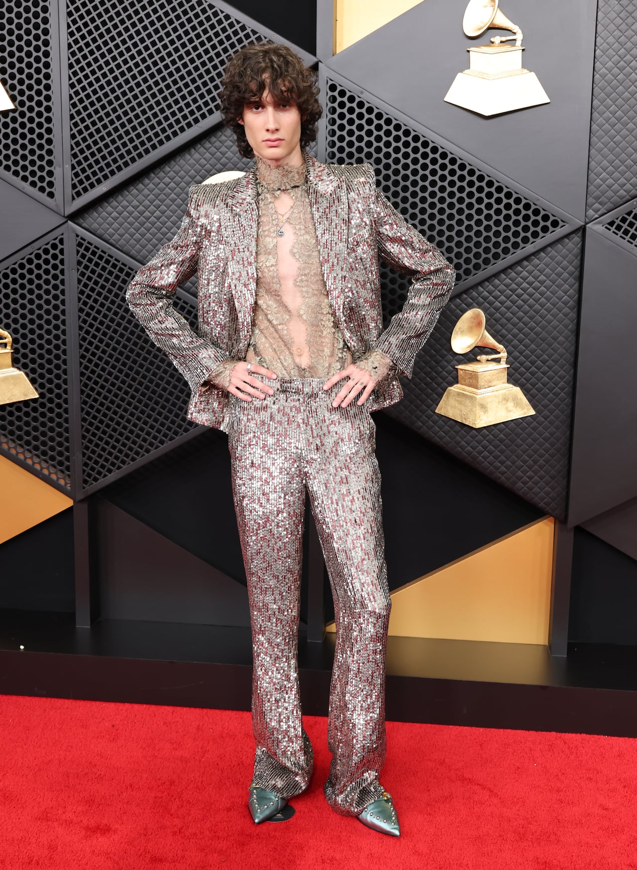 A man poses on the red carpet wearing a shiny silver suit with a sheer lace shirt.
