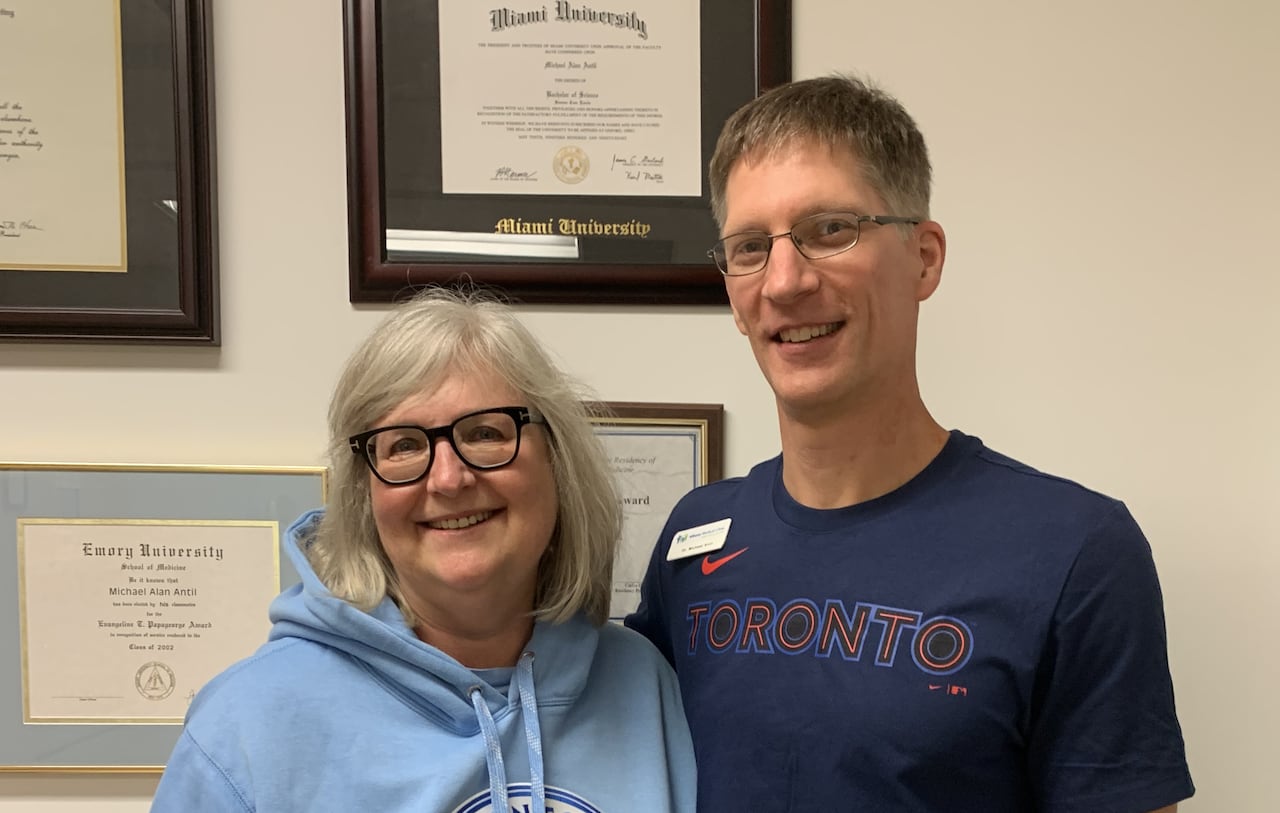 A woman stands with a man, both wearing Toronto t-shirts, in front of a wall with diplomas on it.