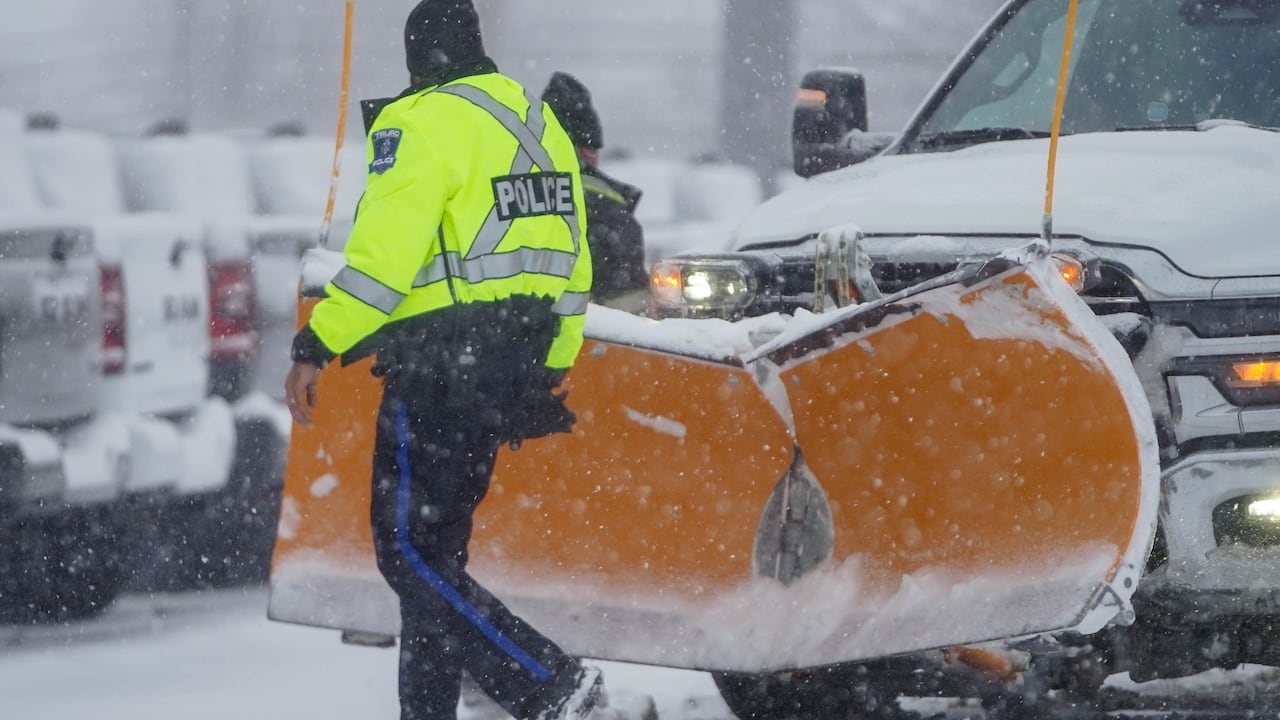 A police officer walks in front of a snowplow as snow falls.