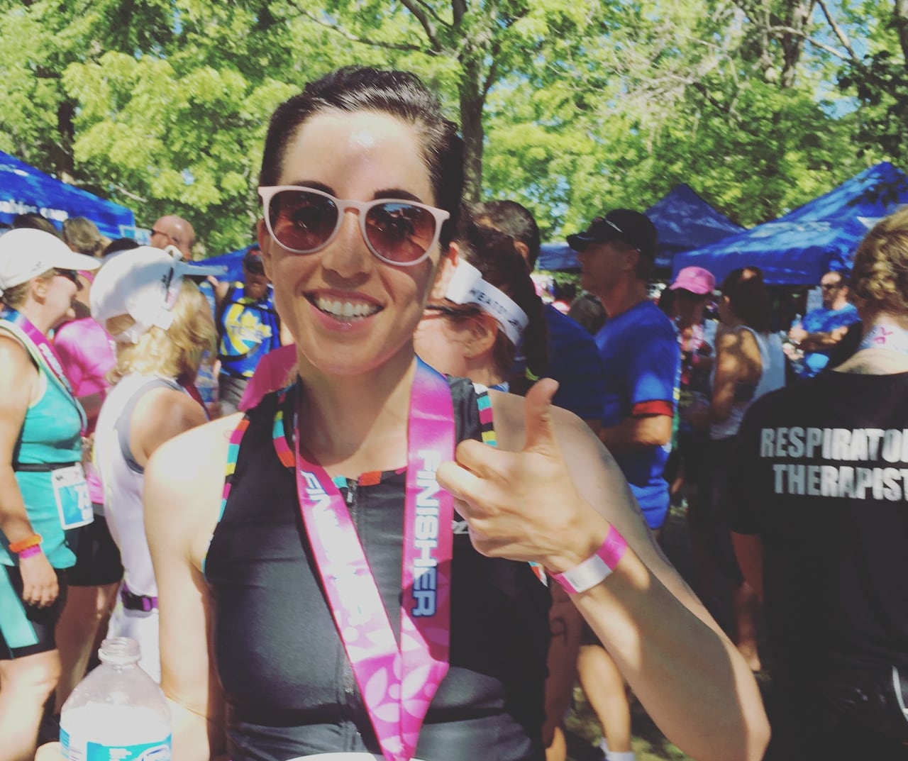 A woman wearing sunglasses and a running bib gives a thumbs up.