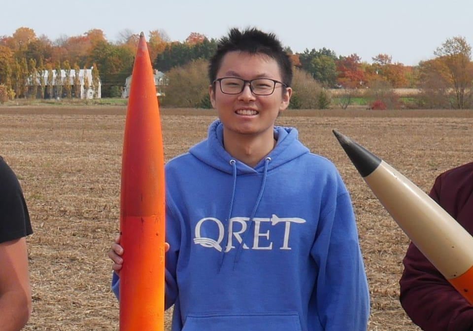 A young man smiling while wearing a hoodie branded with QRET. He's holding a skinny orange rocket