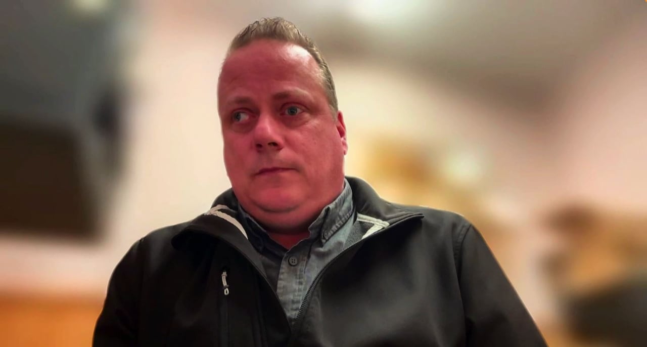 A man wearing a black windbreaker sits. The background is blurred for a Zoom call.