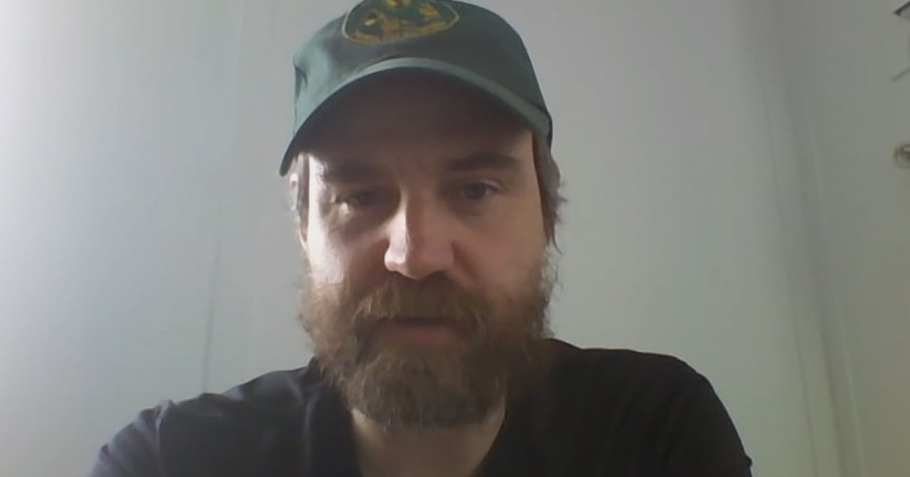 Man is cap speaks in front of a white wall.