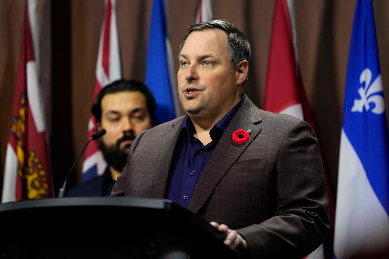 A man in a checkered suit jacket with a poppy on the lapel speaks at a podium in front of various Canadian provincial flags.