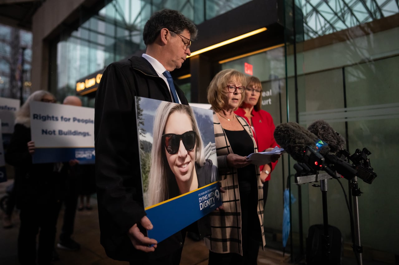 A woman speaks at a podium. Beside her, a man carries a large photo of a young, blond woman in sunglasses