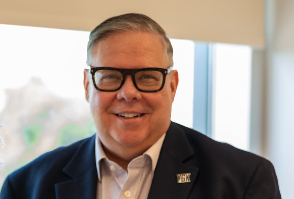 A man and a woman stand together wearing blazers. There is a bright window behind them.