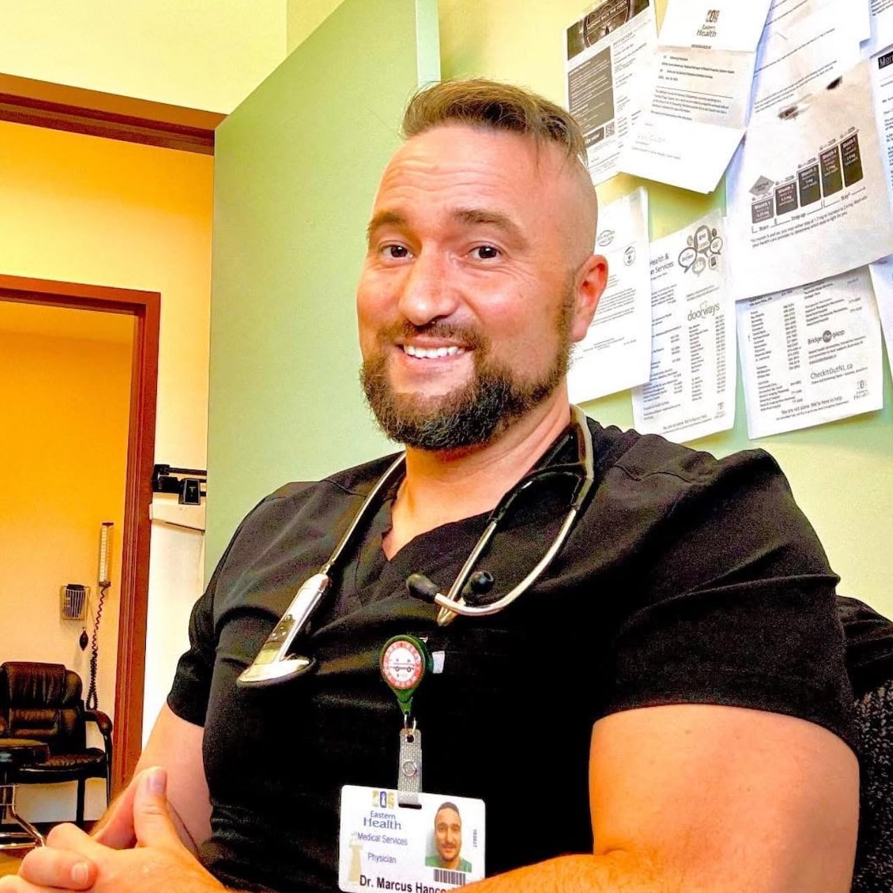A man wearing black scrubs with a stethoscope around his neck.