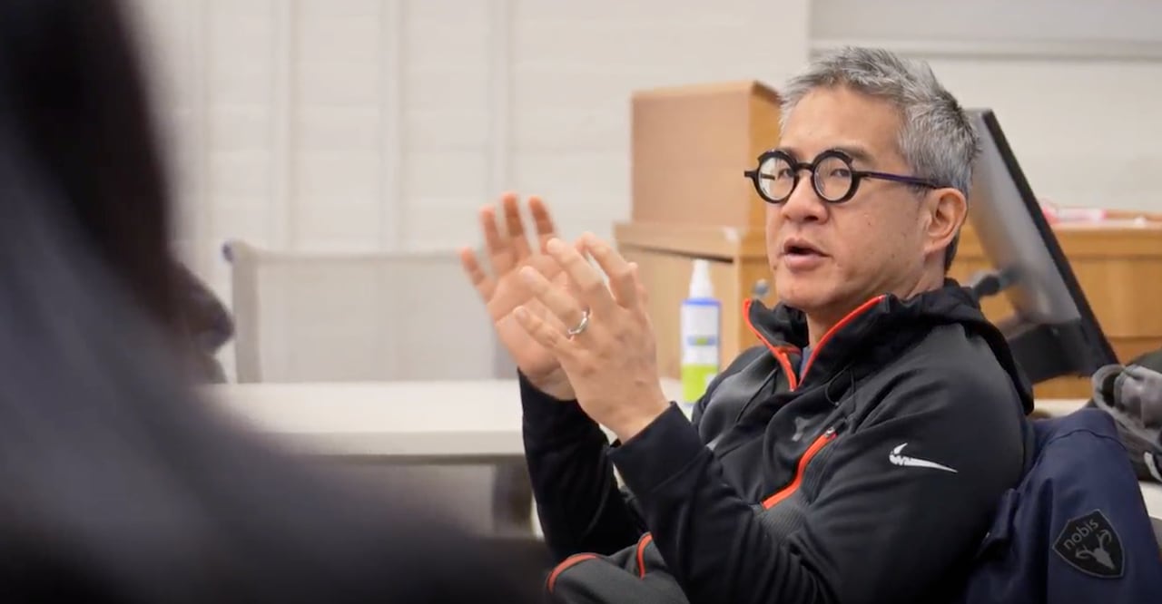 A seated man in glasses and an athletic pullover top gestures with his hands as he speaks to students, one of whom is seen from behind.