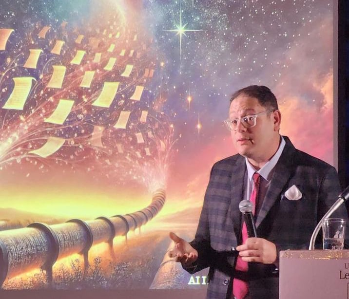 A man speaks to an indoor audience, a projected illustration of papers floating from a pipeline into the night sky displayed behind him.
